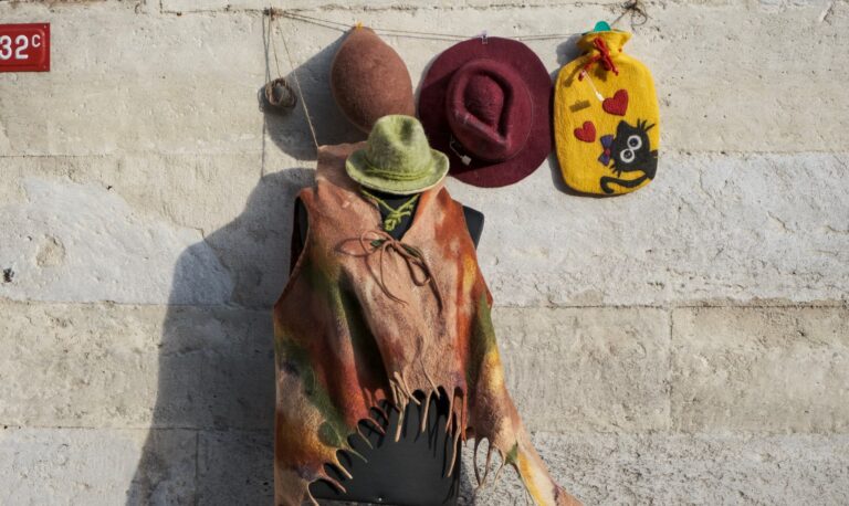 A vibrant display of felt hats and garments hung on a rustic stone wall in sunlight.