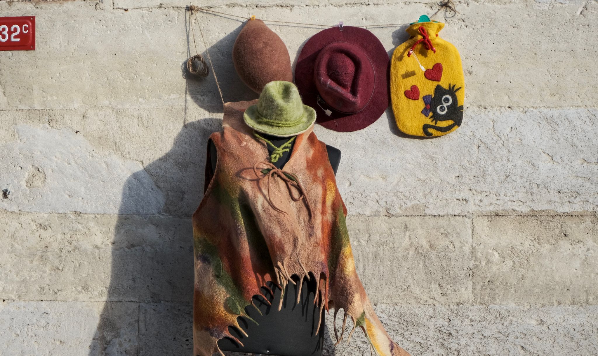 A vibrant display of felt hats and garments hung on a rustic stone wall in sunlight.