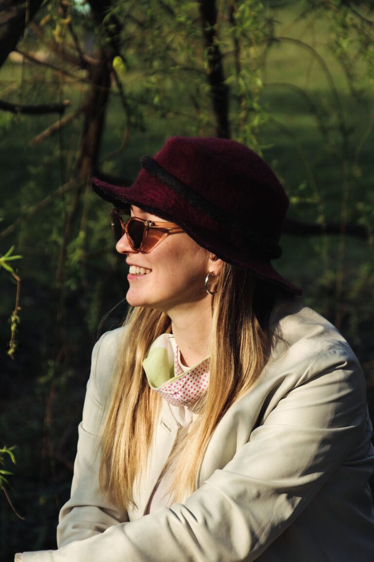 A woman in sunglasses and hat smiling outdoors, embracing the natural setting.